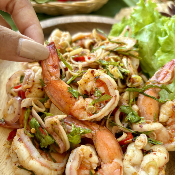 Pla goong salad, Thai shrimp salad, served in a wooden dish with a hand holding a shrimp.