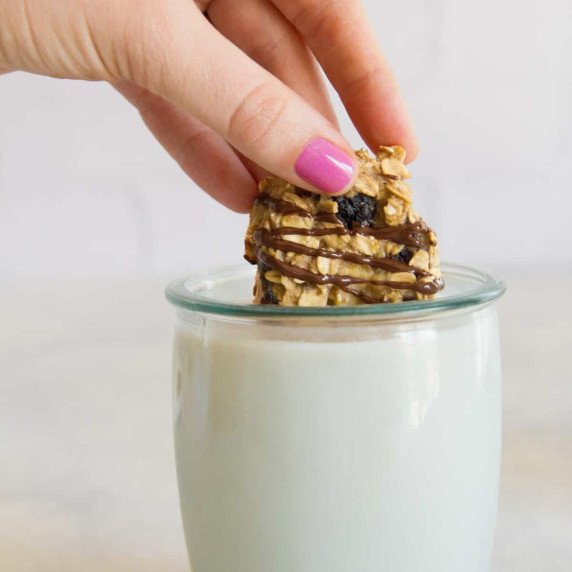 A hand dips an oatmeal raisin snack bite drizzled with chocolate into a glass of milk.