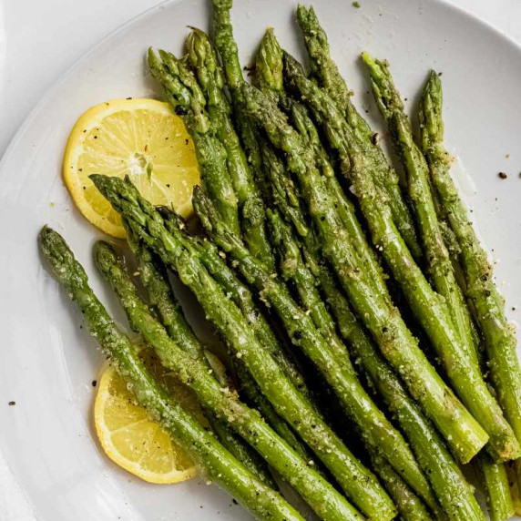 Juicy steamed and seasoned asparagus spears on a white plate with two lemon wheels.