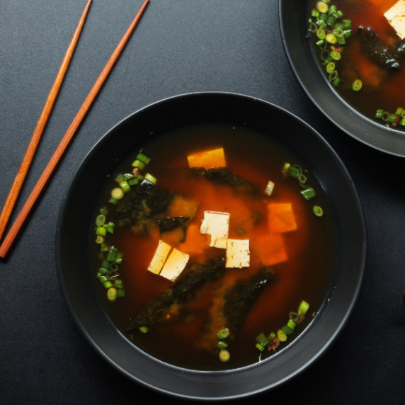A bowl of roasted garlic miso soup in a black bowl on a black surface, with wooden chop sticks.