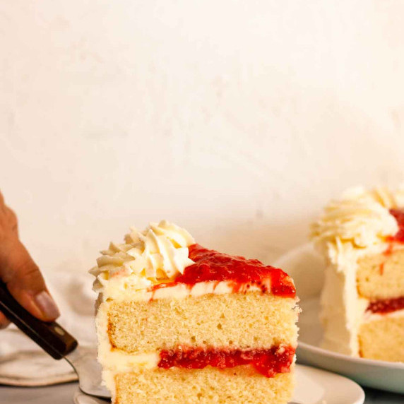 A closeup of a hand placing a slice of vanilla cake with strawberry filling on a white plate.
