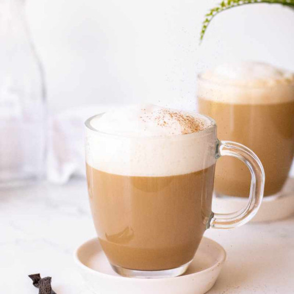 Cinnamon sprinkles down onto the foam atop a homemade vanilla latte in a glass mug.