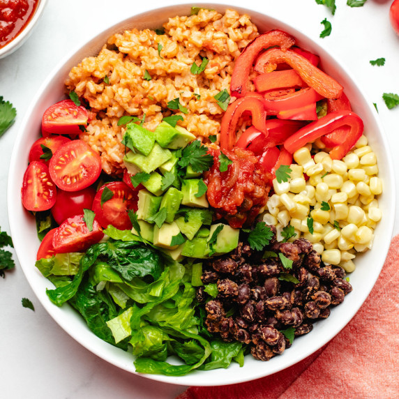 A burrito bowl with beans, corn, rice, red bell peppers, cherry tomatoes, lettuce, and avocado.