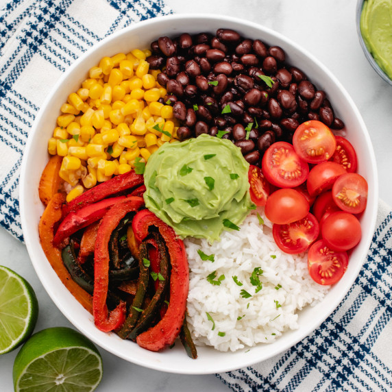 Vegan fajita rice bowl with black beans, cherry tomatoes, corn, cooked peppers, and avocado crema.