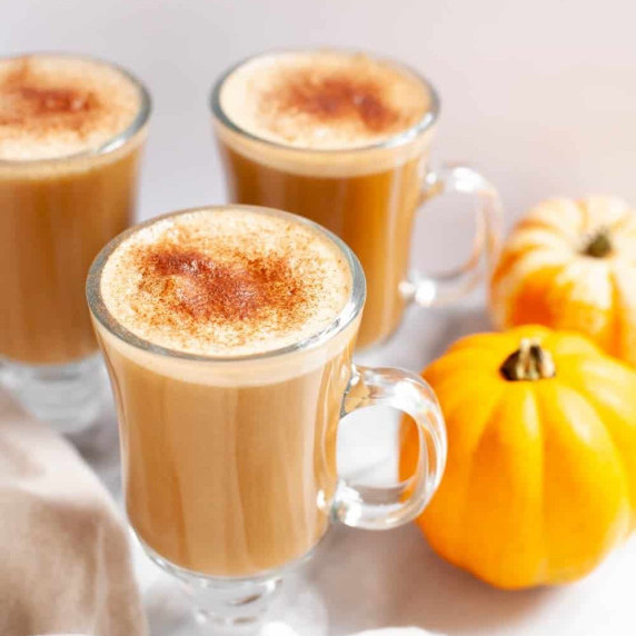 Three homemade pumpkin spice lattes in footed glass mugs stand with two mini pumpkins beside them.