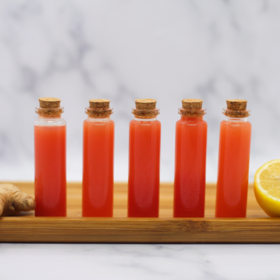 Watermelon Lemon Ginger Juice Wellness Shots in tiny glass vials on a wooden plate.