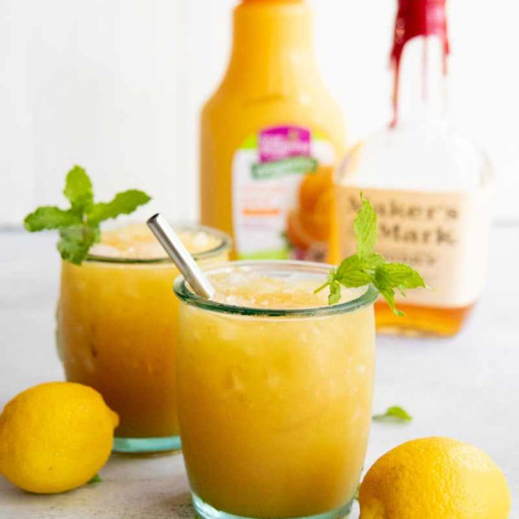 Two glasses of whiskey punch stand with fresh lemons and mint on either side on a white background.