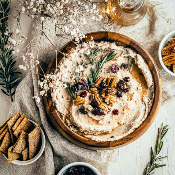 Overhead shot of hummus garnished with pecans, cranberries, and rosemary in a large wooden bowl.