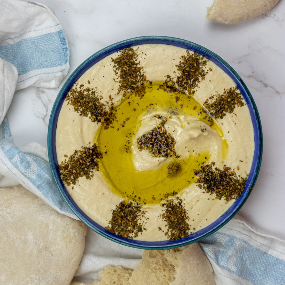 Top down short of hummus in bowl decorated with zaatar. Flanked by pita bread and a tea towl.