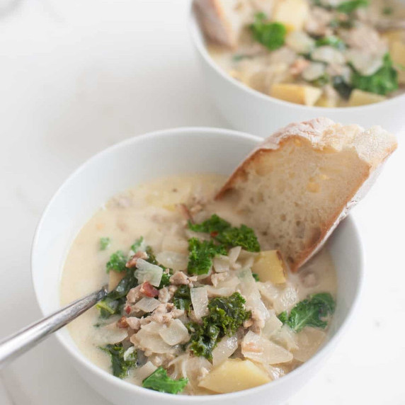 A white bowl filled with zuppa toscana, a spoon, and a piece of bread sits on a white countertop.